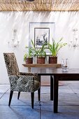 Upholstered, retro-style armchair at wooden table in front of wall with picture flanked by candle sconces on roofed terrace