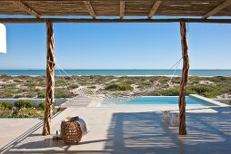 Calm, relaxing atmosphere on sunny, concrete terrace with infinity pool and view of horizon; mesh hammock hanging from two rustic, wooden supports in foreground