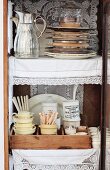 Stacked plates next to thermos jugs, china crockery with vintage-style cutlery and lace doilies in open display cabinet