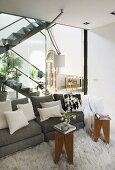 Gray, designer sofa with decorative pillows and small tables on a Flokati rug with a glass wall separating it from the open living room staircase