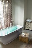 Free-standing bathtub in front of window with closed louver blinds; toiletries on modern side table