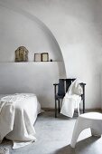 Schwarzer Armlehnenstuhl vor schlicht dekorierter Wandnische und moderner weißer Kunststoffhocker in puristischem Schlafraum