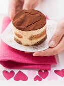 Hands serving an individual tiramisu with a heart drawn in the cocoa powder on top