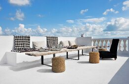 Black and white gingham blankets on masonry bench, rustic wooden table and bast fibre pouffes on seaside terrace