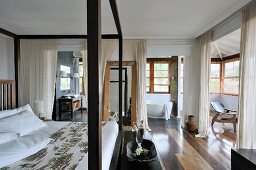 Doppelbett mit Himmelgestell aus dunklem Holz in grossräumigem Schlafzimmer und weiße, bodenlange Vorhänge als Sichtschutz vor dem Bad Ensuite