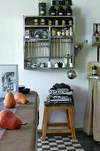 Kitchen utensils, tea caddies and crockery on wall-mounted shelves, tea towels on stool and pears on kitchen table