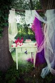 Raspberry cake on cake stand and pink glasses on vintage table between two large tree trunks in garden