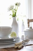 Stacked crockery and cutlery on a table with a vase of white ranunculus