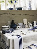 Weiß-blaues Geschirr und Gläser auf weißem Tischtuch ausgestellt an rustikaler Wand unter vergittertem Fenster