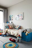 Child's bedroom with toys on rug and blue, cubic pouffe at foot of bed against grey wall