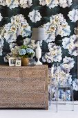 Blumenstrauss und Tischleuchte auf schlichter Holzkommode vor Wand mit weißem Blumenmuster auf dunkler Tapete