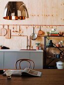 Thonetstuhl an Holztisch, im Hintergrund Küchenzeile unter aufgehängtem Kochgeschirr an Holzwand