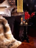 Glamouröses Schlafzimmer mit Felldecken und schwarzem Beistelltischchen, goldfarbenen brennenden Kerzen und roten Orchideenblüten