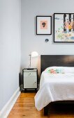 Modern art on wall above black and white bedside cabinet with designer lamp and double bed with dark frame and white bedspread
