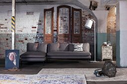 Absurdist arrangement of chaise sofa, standard lamp and rugs against old brick wall