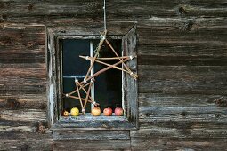 Selbstgebastelte Sterne aus Holzstäben vor adventlichem Fenster mit Äpfeln auf der Fensterbank einer Holzhütte