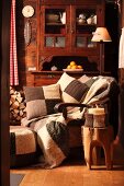 Kerzenglas mit Strick Hülle auf Baumstammhocker vor gemütlichem Armlehnensessel mit gestricken Patchwork Kissen und antikes Buffet an Holzwand