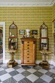 Antique chest of drawers flanked by small marble tables and decorative mirrors on patterned wallpaper in foyer of English country house with marble-tiled floor