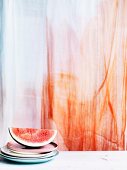 A slice of watermelon on a stack of plates in front of a transparent curtain