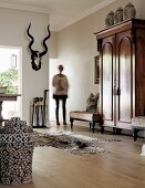 Elegant interior with antique cupboard, colourful rattan basket, zebra-skin rug and wooden floor; woman in background next to hunting trophy on wall