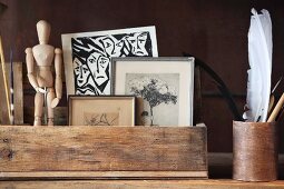 Framed pictured and jointed manikin in wooden crate next to feathers in cylindrical pot