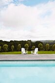 Two white deckchairs next to swimming pool in front of wooded country landscape