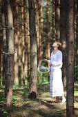 Junge Frau in romantischer, weisser Sommerkleidung und geflochtenem Korb in lichtem Kiefernwald