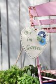 Selbstgefertigter Herzanhänger aus naturfarbenem Filz mit gesticktem Schriftzug und aufgesetzten Stoffrosen, aufgehängt an rostigem Gartenstuhl