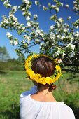 Frau trägt Kranz aus Löwenzahnblüten vor blühendem Apfelbaum