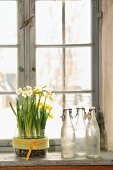 Narcissus in sweet jar next to collection of vintage bottles on windowsill