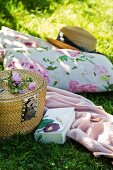 Kissen mit Blumenmuster, Strohhut, Decke, Buch und Picknickkorb auf einer Wiese