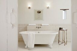 View through open, rustic, white wood sliding door; free-standing, claw-foot bathtub and delicate metal side table against half-height tiled wall