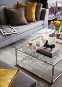 Small sculpture on stacked books and candles on glass and chrome coffee table; sofa, scatter cushions and rug in elegant shades of grey and brown