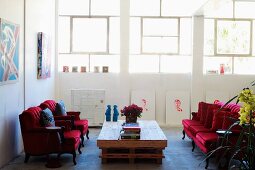 Antique armchairs with red velvet covers around rustic pallet table in loft-style interior with continuous band of windows