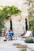 Lesender Mann im Holzliegestuhl in sommerlichem Innenhof mit begrünter hoher Kalksteinmauer eines provenzalischen Stadthauses