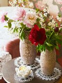 Pfingstrosen in zwei verzierten Vasen und Blütenkerze auf Spitzendeckchen
