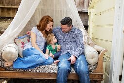 Kleine Tochter zwischen Mutter und Vater gekuschelt auf einem Sofa mit drapiertem Moskitonetz im Freien