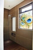 Duschbereich mit Kopfbrause an grossformatiger Fliesenwand und blickdichtes Fenster mit Pfauenfedermotiv