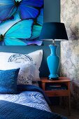 Bed with blankets, pillows and scatter cushions in shades of blue below picture of butterflies; blue bedside lamp on small cabinet