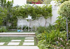Gartenpodest mit filigranen Gartenmöbeln; dahinter eine weiße Gartenmauer mit blau blühenden Bäumchen