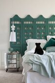 Black dog on bed against wall with antique carved screen painted petrol blue and round bedside table below pendant lamp with white lampshade