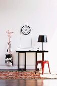 Retro Metall Hocker in Rot vor Wandtisch mit Tischleuchte, an Wand eine Uhr mit Umrisszeichnung eines Turmhauses