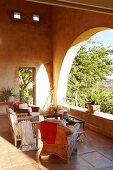 Loggia mit verschiedenen Sesseln und Couchtisch auf Steinfliesenboden und Blick durch Rundbogen in sonnigen Garten