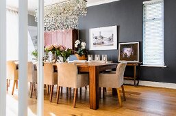 Elegant dining room with anthracite walls, long dining table and upholstered armchairs; pretty sea glass chandelier above table