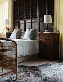 A high double bed in front of antique wood panelling with antique bedside cabinets to either side; a modern angle-poise floor lamp next to each bedside cabinet