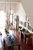 Row of glass spheres on long wooden table, vases of dried flowers and collection of pictures on wall of large foyer