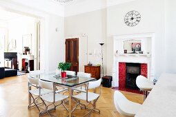 Dining table with glass top and white leather chairs in front of open fireplace in grand interior with open double doors to one side