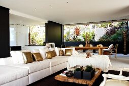 Long sofa with white and golden scatter cushions, low, rustic coffee table and dining area in front of patio with planted beds