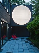 Residential extension with large, white circular area in dark facade at twilight; loungers with red covers on terrace