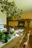 Fresh vegetables on long table in Mediterranean kitchen-dining room; pretty pendant lamp above table and still-life painting on apricot wall